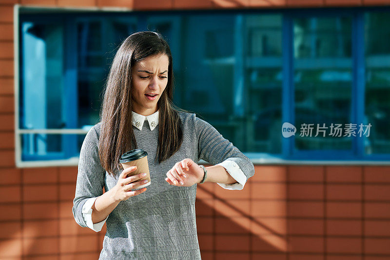 美丽的年轻女子拿着咖啡杯焦急地看着手表对着城市的背景