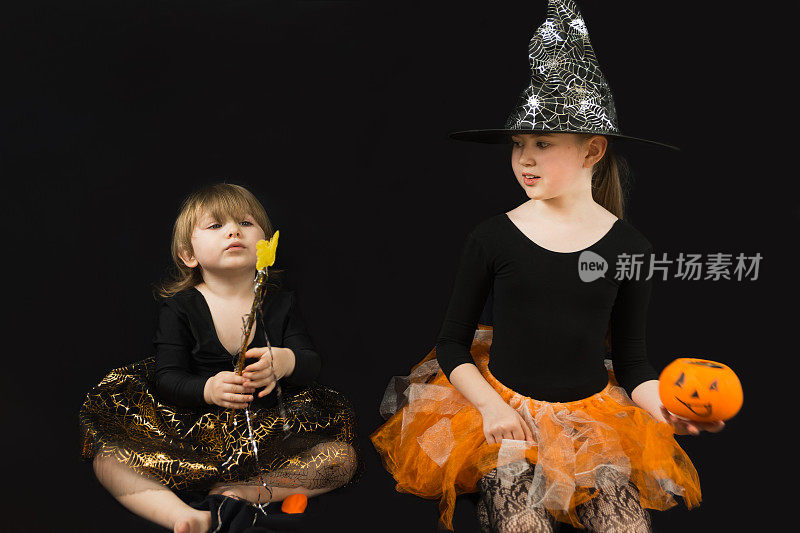 两姐妹装扮成女巫。一件黑色上衣，一顶有蜘蛛网的帽子，一条橙色的裙子。大一点的女孩拿着一个南瓜桶装糖果，小一点的拿着一根魔杖。黑色背景