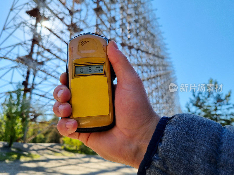 手拿一个黄色的剂量计，显示屏幕上的辐射量。背景是巨大的杜加雷达。温暖的夏日，美丽的平行线