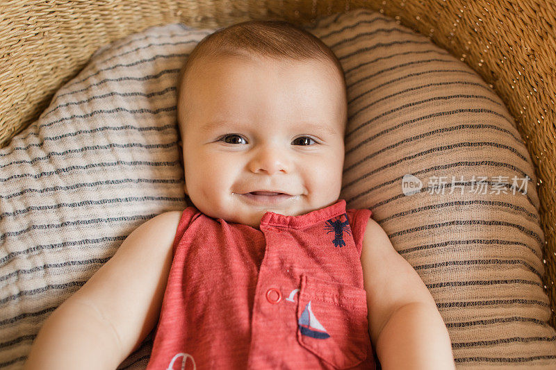 一个18周大的微笑小男孩穿着红色航海服躺在一个舒适的奶油色条纹棉毯在海草摩西篮子