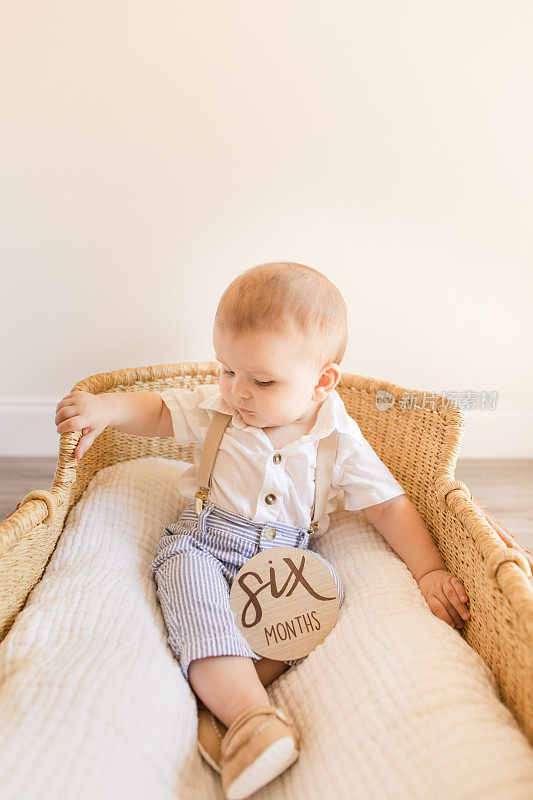 6个月大的小男孩穿着棕色背带的白衬衫，蓝白条纹长裤和棕色乐福鞋，躺在海草摩西篮子里，篮子上有一个木制的圆圈，上面写着“六个月”