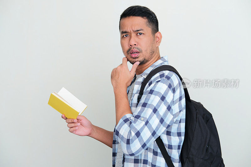 亚洲成年男子背双肩包，拿着一本书，表情困惑