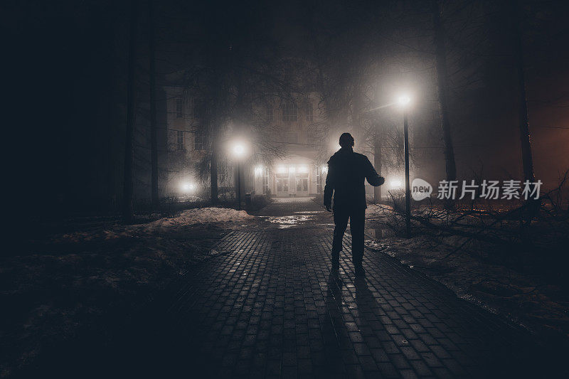 悲伤的人独自走在夜晚雾蒙蒙的公园里的小巷里。后视图