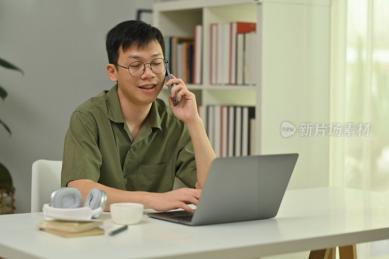 乐观的千禧一代男子戴着眼镜打电话，坐在办公桌前用笔记本电脑。