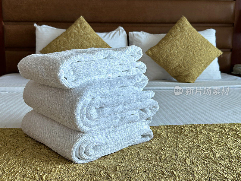 特写图像一堆白色酒店毛巾坐在白色羽绒被床罩上，折叠的浴巾和手巾，酒店房间双人床，一堆白色酒店枕头靠在人皮床头板上，金色靠垫，重点在前景