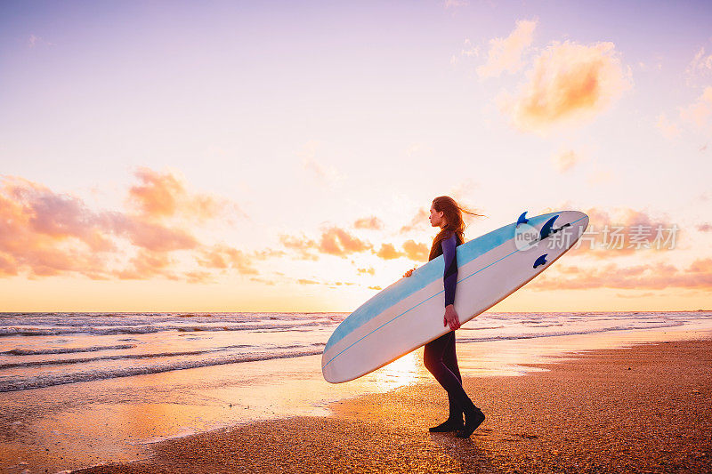 在温暖的日落或日出和海洋中，冲浪女孩拿着冲浪板。