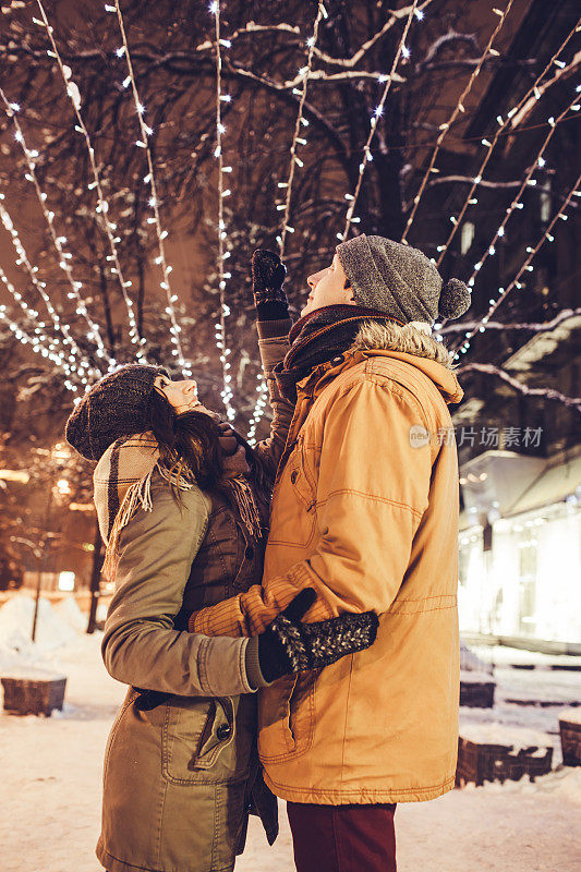 在节日的照明下，一对年轻夫妇在冬季城市中心散步
