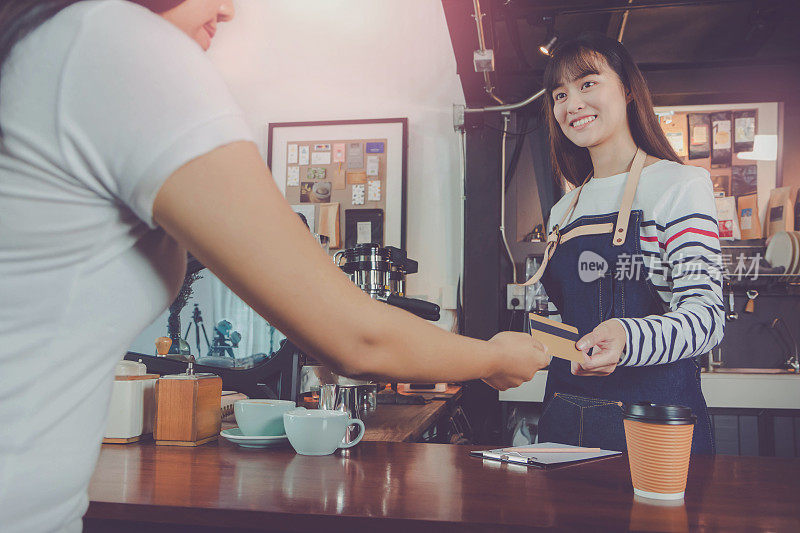 年轻美丽的亚洲女咖啡师穿着蓝色围裙，顾客在咖啡厅柜台用信用卡付款，面带微笑。咖啡店和小型咖啡店的概念。