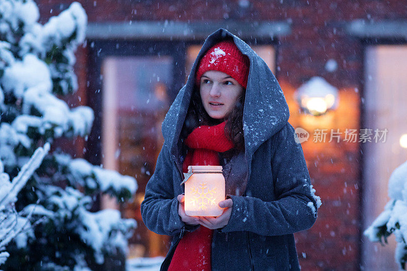 美丽的黑发少女穿着连帽大衣，同时在一个下雪的冬天晚上在户外举着圣诞灯