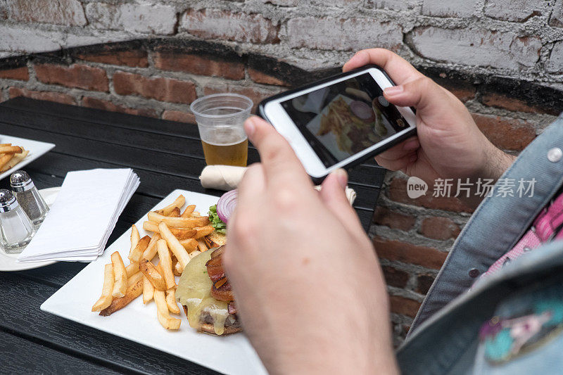 一名男子拿着手机给他的食物拍照。智能手机食品摄影。在户外酒吧用手机给汉堡、啤酒和炸薯条拍照。