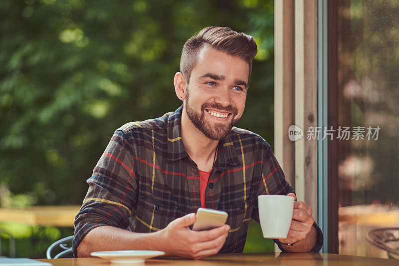 一个帅气的时尚男性，留着时髦的发型和胡子，穿着羊毛衫，喝着咖啡，拿着智能手机在自助餐厅。