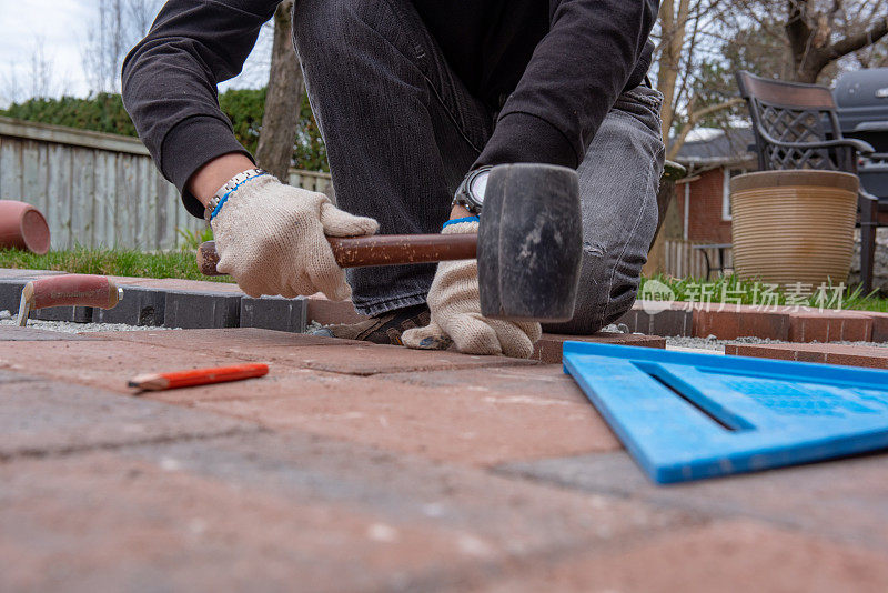 为后院庭院安装铺路砖。一个工人戴着手套的手用锤子来铺石头。