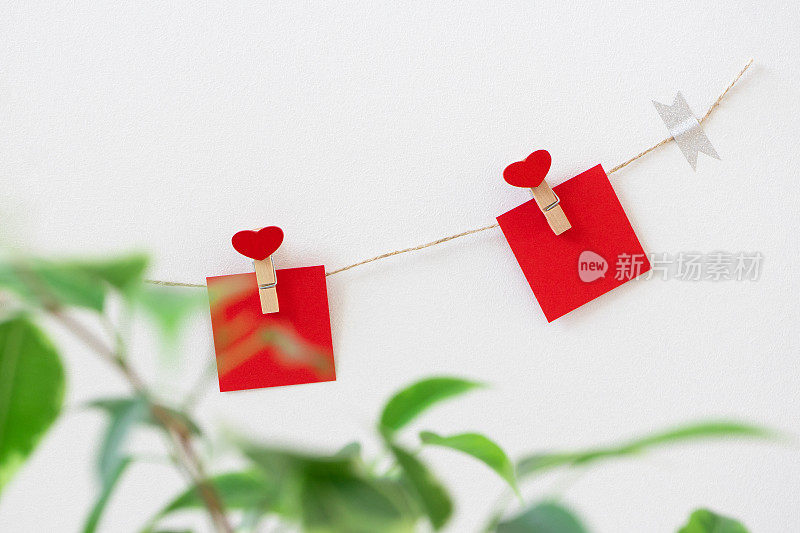 饰有心形衣夹的花环和红纸纸条挂在白色的墙上，横放着。外面是情人节或婚礼派对的装饰品。绿叶散焦