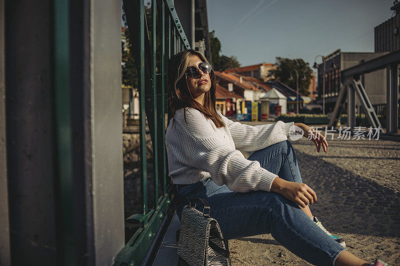 一个时髦的年轻女孩正坐在城市的一座桥上用手机休息