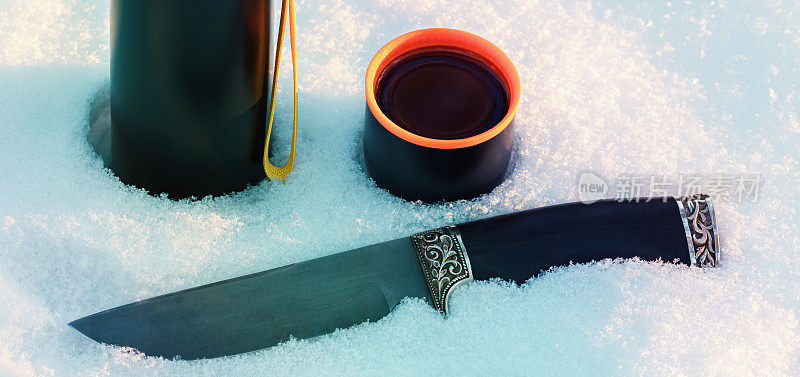 清晨，白雪皑皑的雪地上，热水瓶上放着一杯茶和一把猎刀。冬季狩猎和旅游的概念。