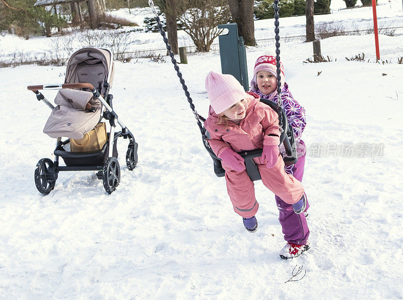 两个白人小女孩，一个3岁，一个5岁，在公园的儿童秋千上荡秋千。春日操场上。第一个春日和阳光。