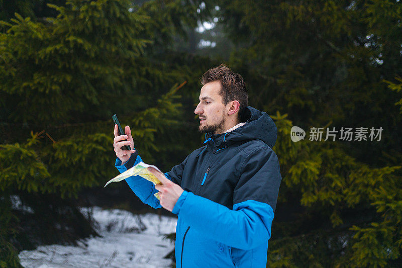 年轻的男性徒步旅行者穿着蓝色夹克，在雾蒙蒙的雪山中迷失了方向，一边看地图，一边看智能手机