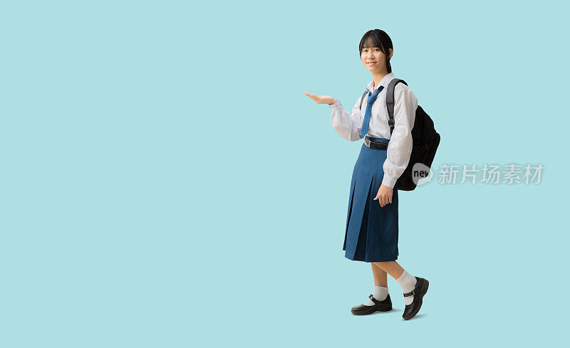 全身亚洲学生女孩穿着制服抱着背包，手拿着敞开的手掌向上留白，在蓝色背景上孤立地用裁剪路径为设计工作留出空的自由空间