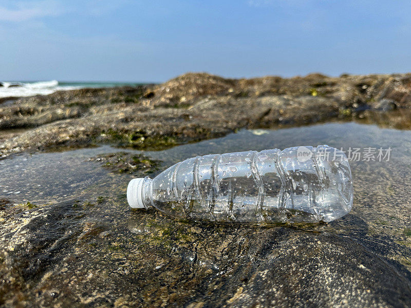 特写图片，空的，透明的塑料水瓶，白色盖子，在退潮时被冲进岩石池，海岸上的海洋垃圾和污染，乱扔的沙子，肮脏的海滩，关注前景，复制空间
