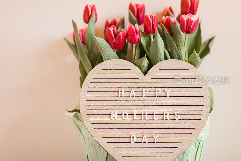 一个心形的字母板，上面写着“母亲节快乐”，明亮的粉红色郁金香盛开，郁郁葱葱的绿色茎和叶子在它后面，在明亮的自然日光下拍摄，中性的米色背景与复制空间