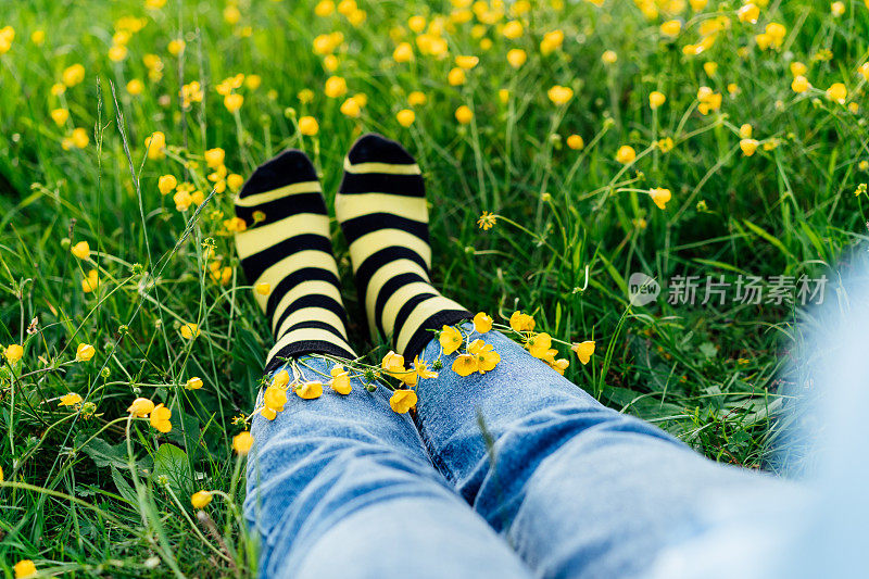 近距离的年轻女性穿着牛仔裤和黑黄相间的条纹袜子，里面有花，坐在盛开的草地上的绿草上。蜜蜂保护理念，开花季节，艺术，创意