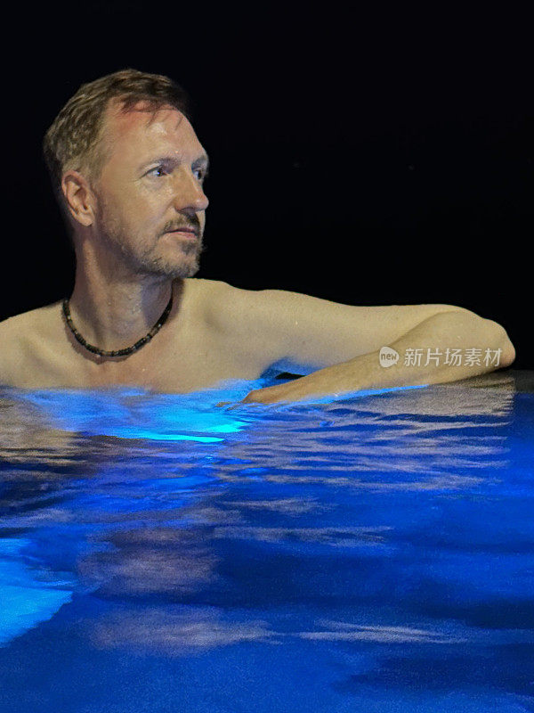 白人男子肩深无边泳池水的特写图像，靠在玻璃墙泳池边，蓝色水下照明，夜空背景，重点在前景