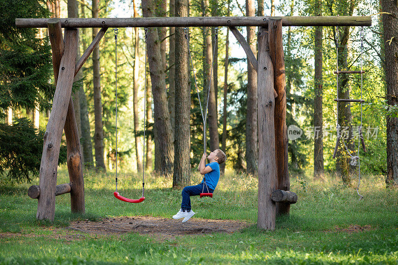在阳光明媚的森林里，一个小男孩坐在一个大秋千上。孩子玩绳子，捻绳子，然后绕着绳子的轴快速旋转