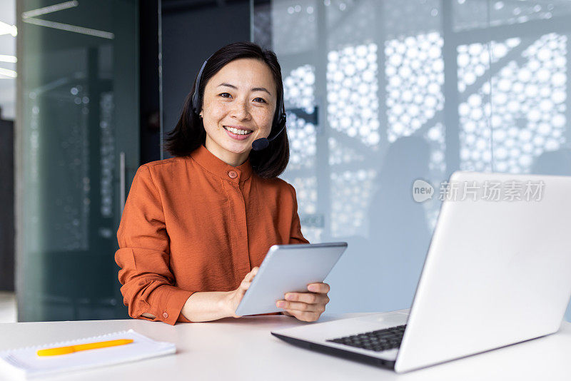 成功满意的女性在线客服支持人员的肖像，亚洲商务女性看着相机，拿着平板电脑，使用耳机和笔记本电脑进行视频通话