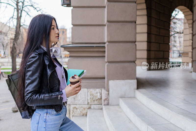 年轻漂亮的女大学生背着双肩包站在大学的楼梯上。她戴着耳机，穿着皮夹克，等待着大学课程的开始。