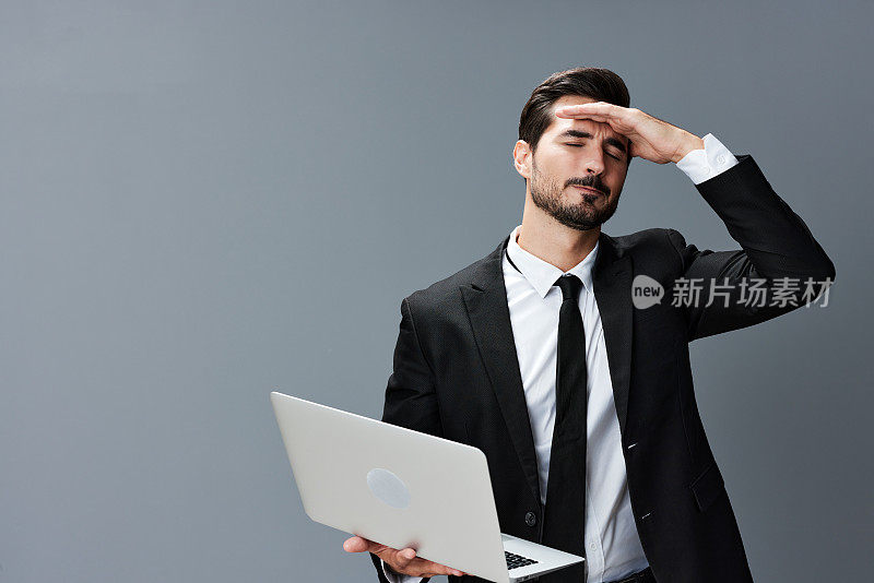 男人有胡子，黑发，商务头痛看着笔记本电脑屏幕上的手在灰色背景的西装。开展网上业务