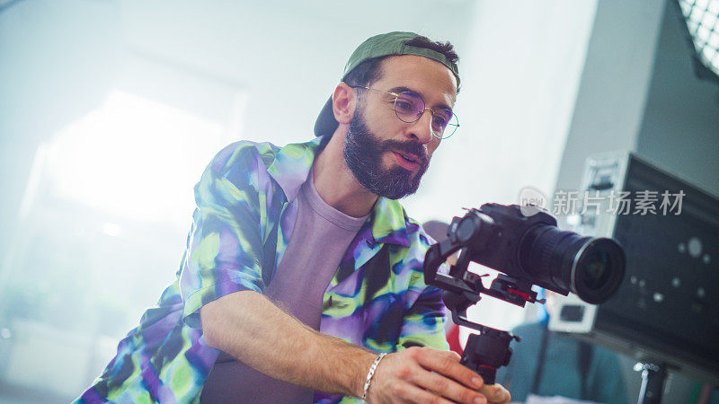 有胡子的年轻男摄影师，穿着彩色夹克，戴着眼镜，在室内电影拍摄中调整现代相机的设置。电影制作中的创造力和专业精神。