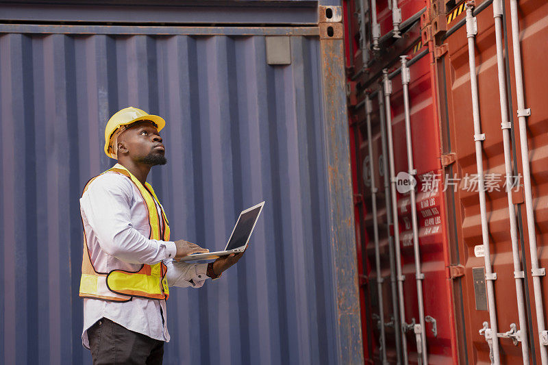 非洲黑人男子戴安全帽，身穿背心，手持笔记本电脑检查集装箱货物上的电子封条，年轻的男性技术人员在船坞工作