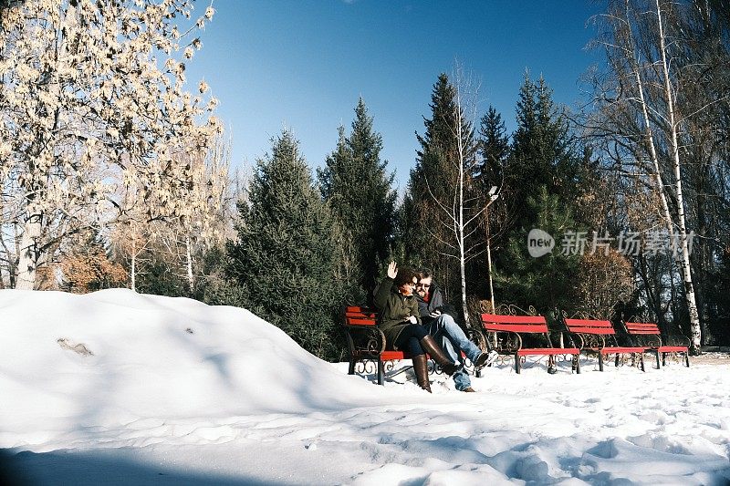 一对成熟的夫妇坐在冬天公园的长椅上使用手机