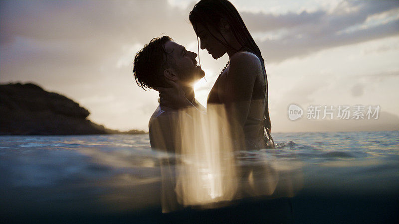 浪漫的异性情侣在美丽的金色夕阳下在海里游泳。在水里嬉戏拥抱