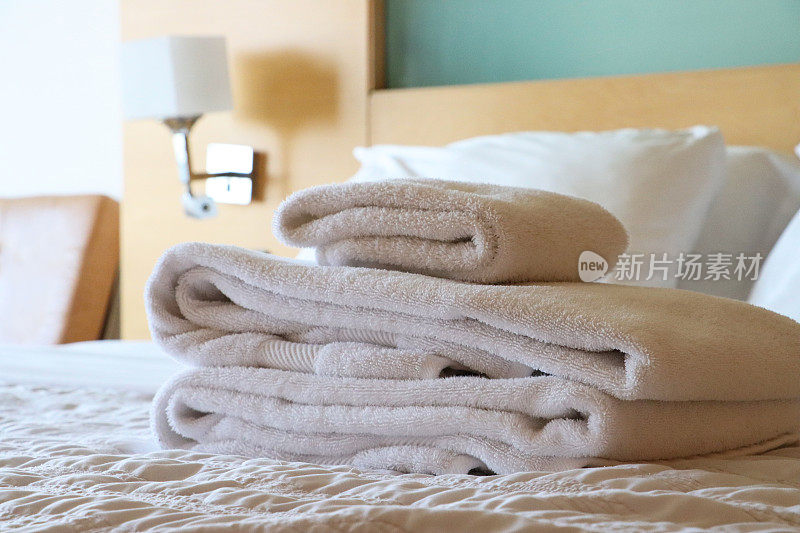 特写图像的堆叠白色酒店毛巾坐在白色羽绒被床罩，折叠浴床单和手巾，枕头靠在木床头板，床头柜，壁灯，皮椅，重点在前景