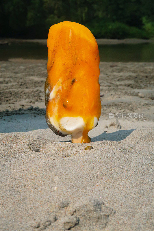 近距离图像融化的冰淇淋棒棒糖与木棒棒棒卡在沙滩上的沙子，橙色的冰棒在退潮时在海水的边缘，芒果和橙色泥覆盖的冰淇淋pop反对海岸背景，重点在前景