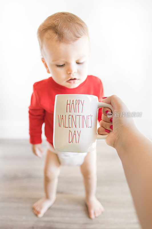 一个14个月大的可爱男婴光着脚，穿着写着“最小的情人节”的红毛衣和布尿布，坐在木制硬木地板上，他的妈妈在他面前举着一个写着“情人节快乐”的杯子