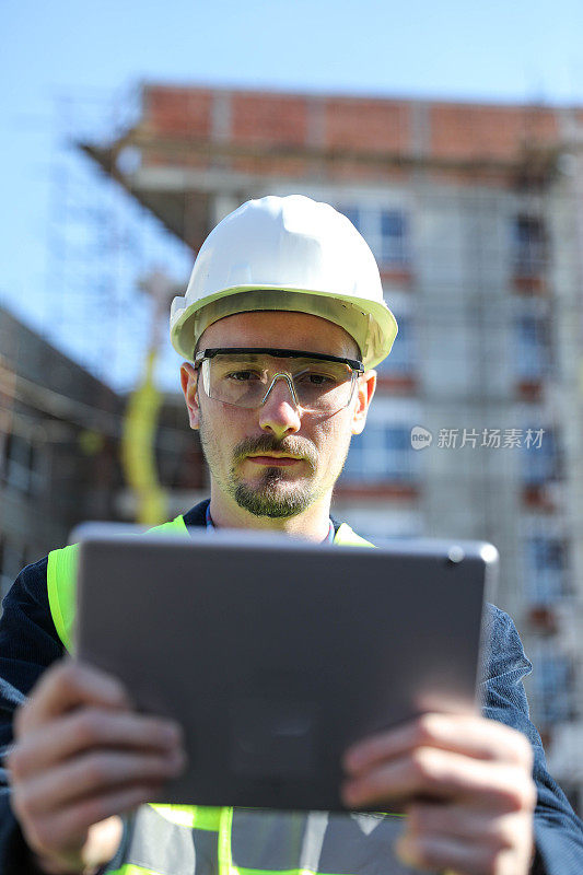 男建筑师在工地使用数码平板电脑，他穿着防护背心和头盔