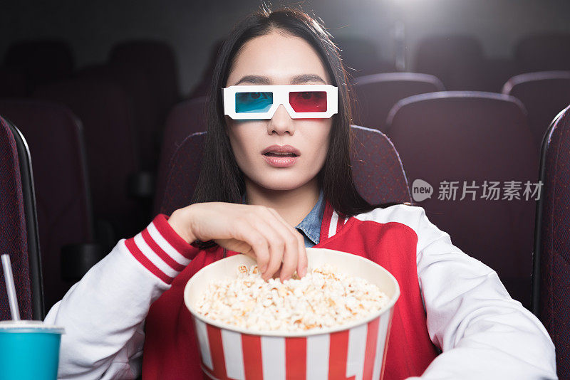 年轻的亚洲女孩戴着3d眼镜，拿着一大篮爆米花在电影院看电影