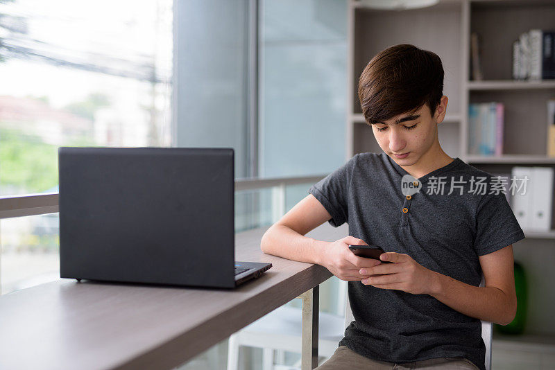 年轻英俊的伊朗少年在家里放松时使用笔记本电脑和手机的肖像