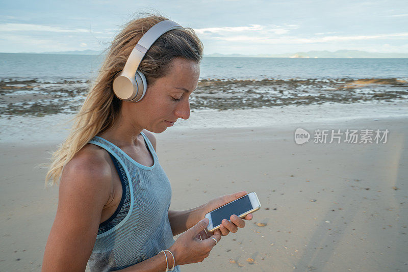 运动的年轻女子走在海滩上的手机和耳机享受早晨日出在海边的自然