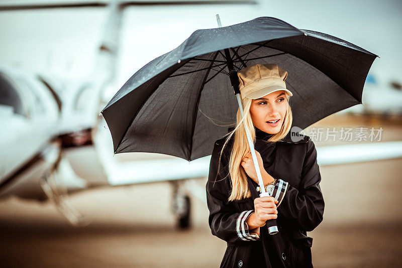 一个漂亮的金发女人，撑着一把黑伞，站在一架停在机场停机坪上的私人飞机前