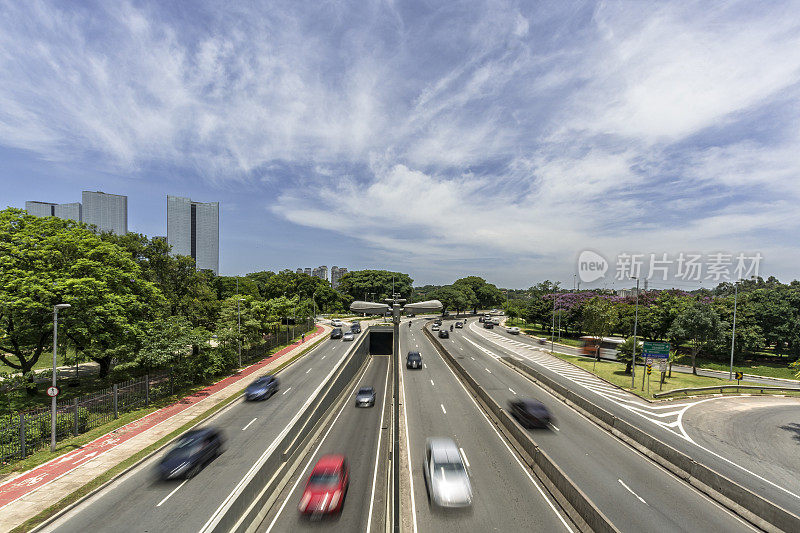 宽阔的四车道高速公路，车水马龙，两边都是树木，蓝天白云