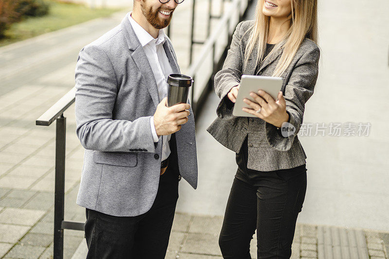 近距离观察男性经理和女性员工站在办公大楼前讨论人事问题，经理喝咖啡和员工使用数字平板电脑