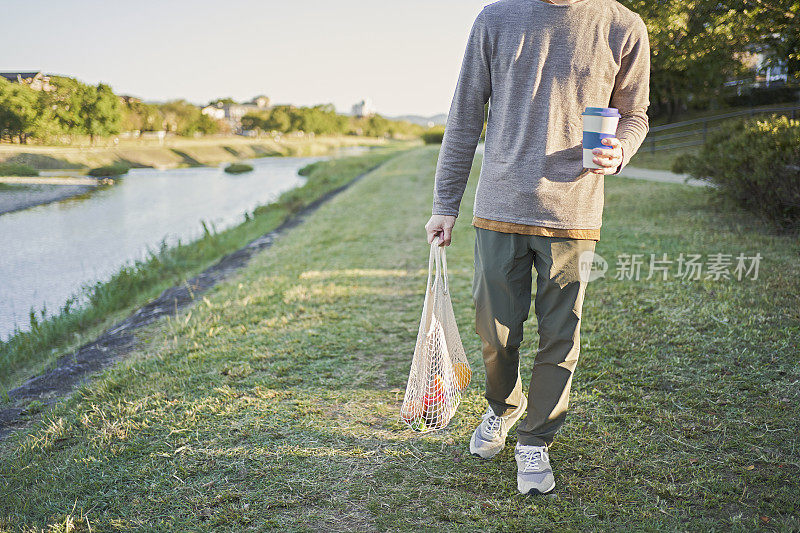 一名日本男子背着装在网眼袋里的杂货，沿着河边的木板路行走