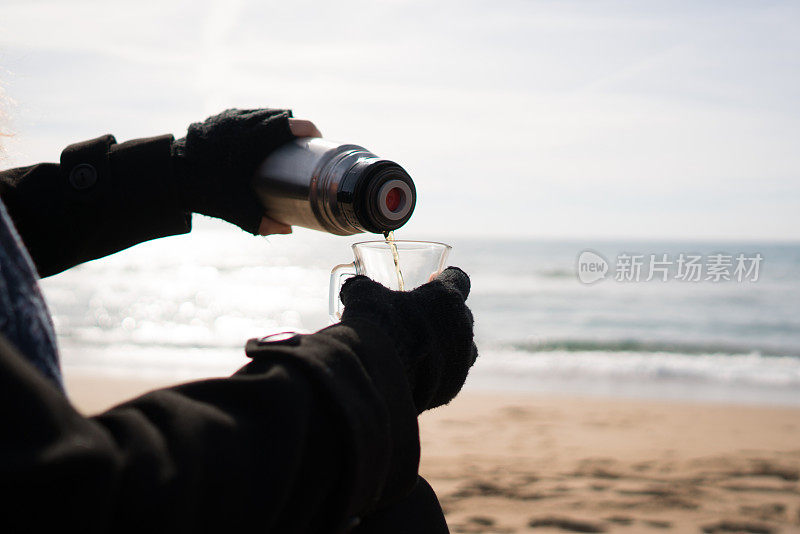 冬天，在海滩上，一名妇女正在把热水瓶里的茶倒进马克杯里。