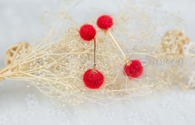 红色的绒球耳环的形状像两个毛茸茸的球。耳环旁边有藤制品和花束样品。这些东西都放在薄纱上。这耳环看起来很漂亮，很时尚。