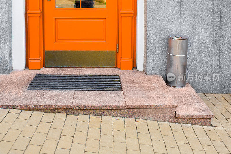 带脚垫的花岗岩门槛，靠近橙色木质前门和铁垃圾桶，靠近建筑立面，灰色覆层和黄色石瓦路面特写，没有人。
