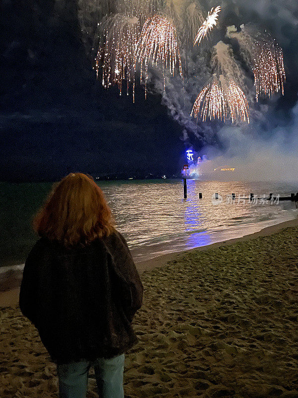 年轻男子的剪影，站在海滩的水边，观看烟花表演，烟雾和夜空中的火花