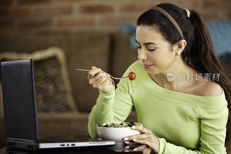 特写的西班牙裔可爱的年轻女子吃素食沙拉在午餐时使用笔记本电脑